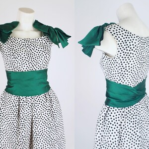 K&ouml;nnte beinhalten: Vintage Kleid mit wei&szlig;em Hintergrund und schwarzen sternf&ouml;rmigen Mustern. Das Kleid hat einen gr&uuml;nen Satin-Sch&auml;rpe und Schulterakzente. Das Kleid wird auf einer Schaufensterpuppe pr&auml;sentiert.