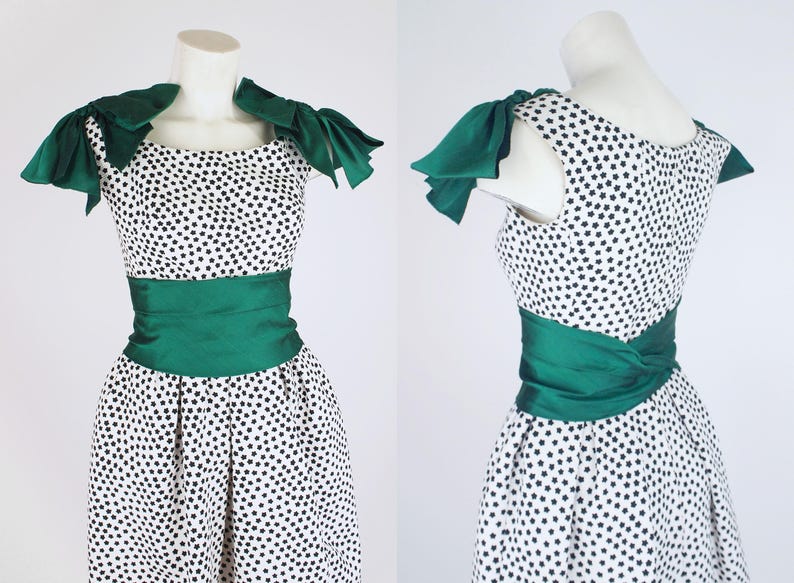 K&ouml;nnte beinhalten: Vintage Kleid mit wei&szlig;em Hintergrund und schwarzen sternf&ouml;rmigen Mustern. Das Kleid hat einen gr&uuml;nen Satin-Sch&auml;rpe und Schulterakzente. Das Kleid wird auf einer Schaufensterpuppe pr&auml;sentiert.