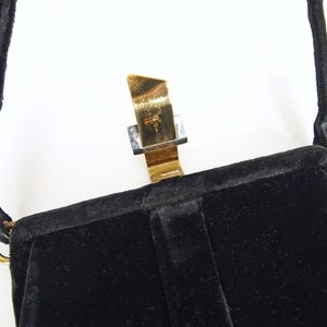 Puede incluir: Un bolso de noche de terciopelo negro con un cierre de metal dorado. El bolso tiene una sola correa.
