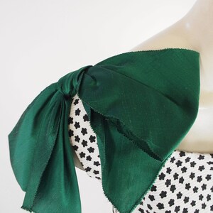 K&ouml;nnte beinhalten: Nahaufnahme eines gr&uuml;nen Stoffschleifen-Details an einem Kleidungsst&uuml;ck. Die Schleife ist geknotet und &uuml;ber einem Stoff mit schwarz-wei&szlig;em Blumenmuster drapiert. Der Stoff hat ein strukturiertes Aussehen.