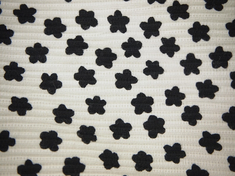 K&ouml;nnte beinhalten: Nahaufnahme eines wei&szlig;en Stoffes mit einem sich wiederholenden Muster aus schwarzen, f&uuml;nfbl&auml;ttrigen Bl&uuml;tenformen. Die Blumen sind gleichm&auml;&szlig;ig verteilt und erzeugen ein strukturiertes Design. Der Hintergrund hat eine subtile, gerippte Textur.
