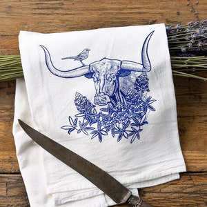 Puede incluir: Un pa&ntilde;o de cocina de algod&oacute;n blanco con una ilustraci&oacute;n en tinta azul de una cabeza de buey longhorn con un peque&ntilde;o p&aacute;jaro posado en un cuerno, rodeado de flores. Un cuchillo de metal con mango texturizado descansa sobre la tela doblada.