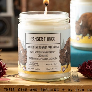 May include: A lit 8 oz. soy wax candle in a clear glass jar with a label reading RANGER THINGS, SMELLS LIKE TOURIST FREE PARKS, WITH NOTES OF WARM EARTHY CEDAR, AND BASE NOTES OF VANILLA AND MUSK, featuring a brown bison illustration.