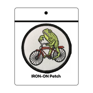 May include: A circular iron-on patch featuring an embroidered green toad riding a red bicycle. The patch has a white background and a thick black stitched border. The text IRON-ON Patch is printed in black at the bottom of the white packaging card.