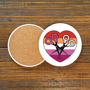 May include: Two round ceramic coasters. One coaster features a heart design in the colors of the lesbian pride flag, with scissors. The other coaster has a cork base. The coasters are on a wooden surface.