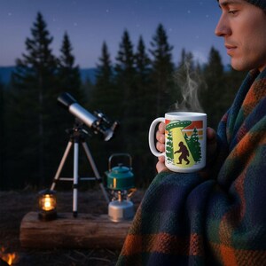 A person is sitting outdoors at night, wrapped in a blanket and holding a mug with a picture of a Sasquatch on it. In the background, there is a telescope and a lantern, suggesting a camping or outdoor activity setting.