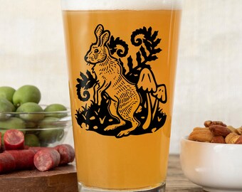 May include: A clear glass pint glass filled with a golden-colored beverage, featuring a black graphic print of a standing hare surrounded by stylized ferns and two mushrooms on the front surface of the glass.