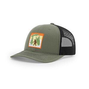 Op de afbeelding: Een olijfgroene truckerpet met een zwarte mesh achterkant en een rechthoekige patch op de voorkant. De patch toont een afbeelding van een bruin Bigfoot-silhouet in een bos onder een groene UFO met een gele lichtstraal en een oranje rand.