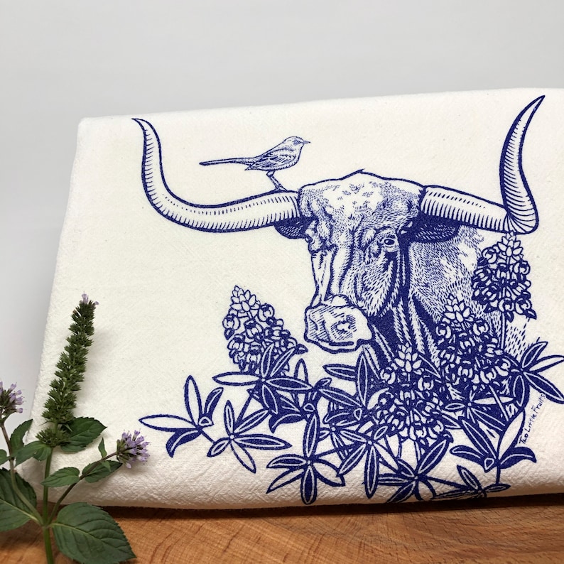Puede incluir: Un pa&ntilde;o de cocina de tela blanca con una ilustraci&oacute;n en tinta azul de un buey longhorn con un peque&ntilde;o p&aacute;jaro posado en un cuerno, rodeado de flores de lupino azules. El texto Two Little Fruits est&aacute; impreso en letras azules abajo a la derecha.