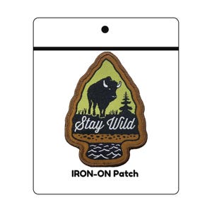 May include: An iron-on patch with a bison and pine tree design, set against a green background. The patch is shaped like an arrowhead and features the words "Stay Wild" in white script. The bottom of the patch has a water wave design.
