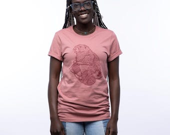 May include: A dusty rose short-sleeved t-shirt featuring a dark maroon line art graphic of a groundhog holding a ball of yarn. The shirt has a crew neckline and cuffed sleeves, providing a casual look with a detailed animal illustration.
