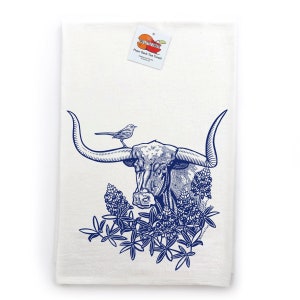 Puede incluir: Un pa&ntilde;o de cocina blanco con una ilustraci&oacute;n en tinta azul de un buey longhorn con un peque&ntilde;o p&aacute;jaro posado en un cuerno, rodeado de flores de lupino. Una etiqueta en la parte superior muestra el texto Two Little Fruits Flour Sack Tea Towel.