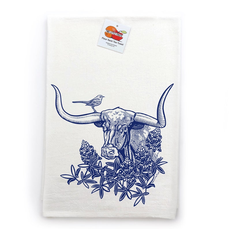 Puede incluir: Un pa&ntilde;o de cocina blanco con una ilustraci&oacute;n en tinta azul de un buey longhorn con un peque&ntilde;o p&aacute;jaro posado en un cuerno, rodeado de flores de lupino. Una etiqueta en la parte superior muestra el texto Two Little Fruits Flour Sack Tea Towel.