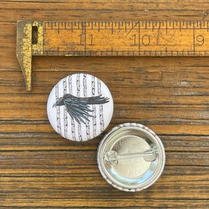 May include: A circular pinback button featuring a black raven illustration against a white background with vertical gray lines, positioned next to a metal ruler showing a diameter of approximately 1.25 inches for the button.