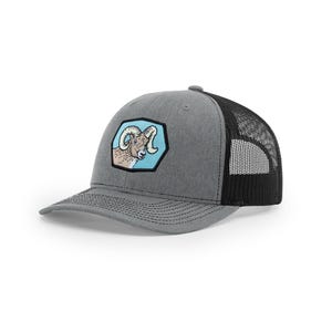 May include: A gray heather baseball cap with a black mesh back and a structured front panel. The front features a hexagonal patch with a light blue background and an illustration of a bighorn sheep head with curved horns and tan fur.