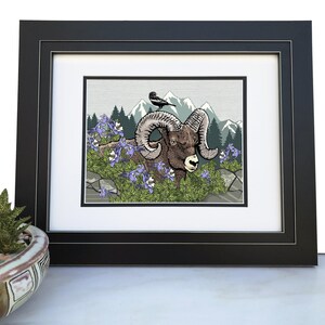 May include: A framed illustration of a bighorn sheep with large curled horns in a mountain landscape. The sheep is brown and white with a black nose. The sheep is surrounded by green foliage and purple flowers. A small black bird is perched on a rock behind the sheep. The background is a gray and green mountain scene.