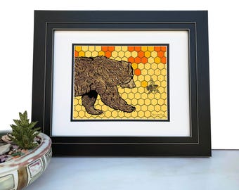 May include: A framed art print featuring a brown bear walking toward a small bee against a yellow and orange honeycomb background. The print is enclosed in a black frame with a white mat. The text "The Little Print" is visible in the bottom right corner.