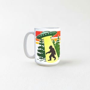 A mug with a graphic design depicting an alien spacecraft hovering over a silhouetted figure walking through a forest.