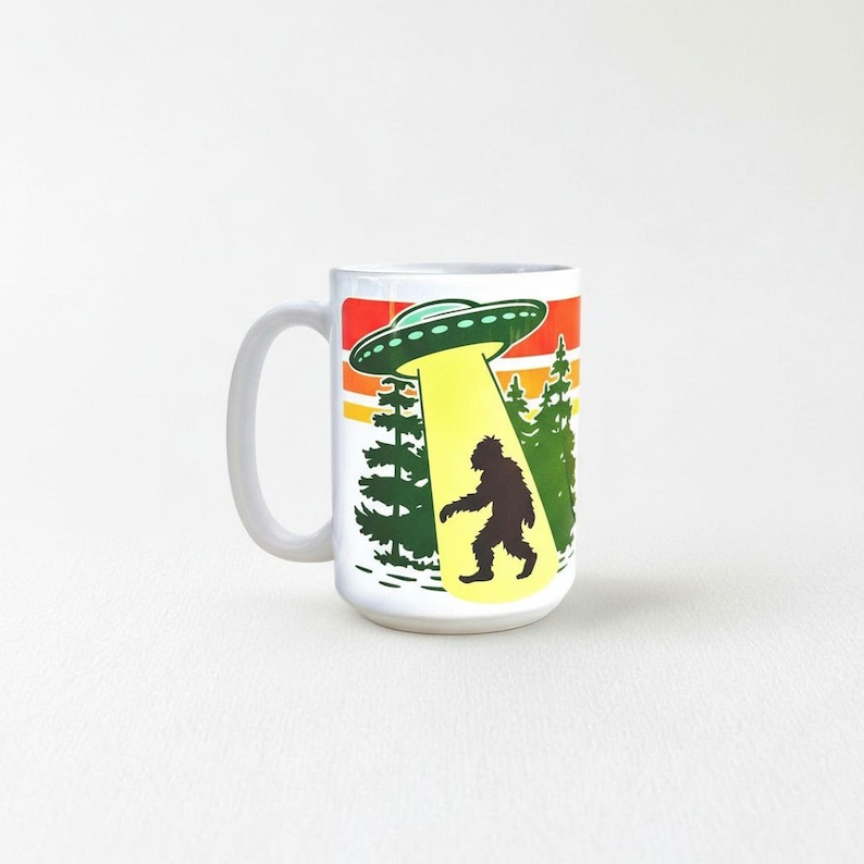 A mug with a graphic design depicting an alien spacecraft hovering over a silhouetted figure walking through a forest.