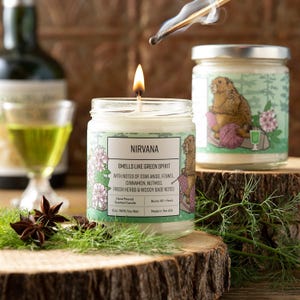May include: A clear glass jar candle with a white label featuring a groundhog illustration and the text NIRVANA SMELLS LIKE GREEN SPIRIT. The label lists notes of star anise, fennel, cinnamon, nutmeg, fresh herbs, and woody base notes.