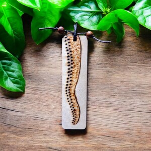 May include: A wooden bookmark featuring a black and brown design of a wavy line with dots. The bookmark is on a wooden surface with green leaves in the background.