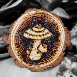 May include: A wood slice with a woodburned image of a mushroom with a light brown cap and a white stem. The wood slice has a natural wood grain and a dark brown border.