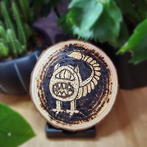 May include: A wooden coaster with a burnt design of a creature with a large eye and a mouth. The creature has a wing-like structure on its back.