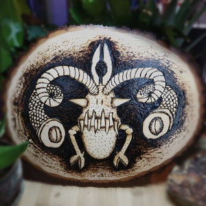 May include: A wood-burned design of a creature with horns and a toothy grin on a round wooden slice.