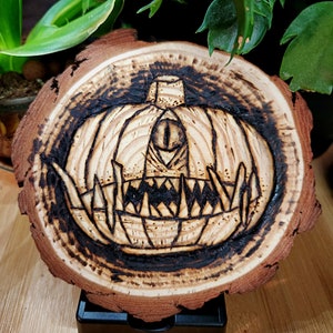 May include: A wooden slice with a woodburned image of a pumpkin with a single eye and sharp teeth. The pumpkin is dark brown and the background is light brown.