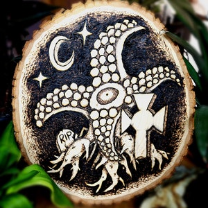 May include: A wood slice with a black and white design of a creature with tentacles and a cross. The creature has a large eye and is surrounded by stars and a crescent moon. The words "RIP" are carved into the wood.
