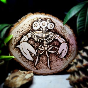 May include: A wood slice with a pyrography design of a skeletal crab-like creature with three eyes. The artwork is burned into the light-colored wood, with dark brown detailing. The crab has large claws and a rib cage. The wood slice has a natural, rustic edge.