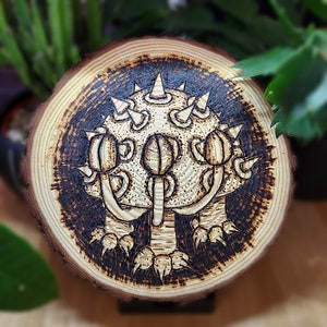 Puede incluir: Una rodaja redonda de madera con un diseño quemado de una criatura con múltiples ojos y espinas. La criatura tiene un cuerpo redondo con tres patas y tres ojos.