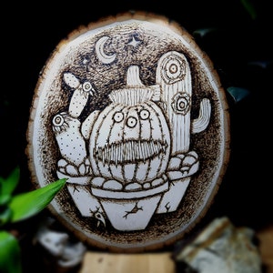 May include: A woodburned design on a wooden slice featuring a cactus with a toothy grin, two smaller cacti, and a crescent moon and star.
