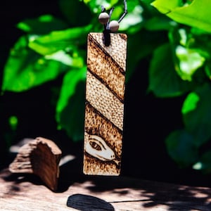May include: A wooden bookmark with a burned design of a snake's head and scales. The bookmark is hanging from a black cord with white beads.