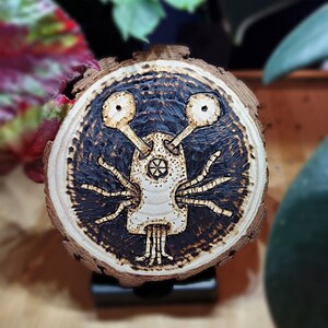 Puede incluir: Una rodaja de madera con un diseño de madera quemada en negro y blanco de una criatura con ojos grandes, un cuerpo redondo y múltiples patas.