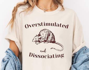 Pode incluir: Uma camiseta branca com uma ilustração de rato marrom e o texto "Overstimulated and Dissociating".