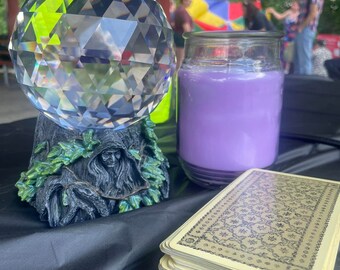 May include: A crystal ball sits on a green and brown figurine of a tree spirit. A purple candle and a stack of tarot cards are on a black surface.