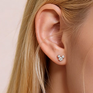 Könnte beinhalten: Nahaufnahme eines Ohrs mit einem kleinen Ohrstecker aus Silber. Der Ohrring hat die Form eines Mäusekopfes mit drei runden, klaren Edelsteinen. Die Person hat lange, blonde Haare.