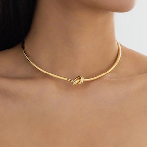 Könnte beinhalten: Goldfarbene Halskette im Choker-Stil mit Knoten-Design. Die schlichte, elegante Halskette hat einen Knoten in der Mitte. Ein stilvolles Schmuckstück.