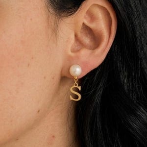 Könnte beinhalten: Ein goldfarbener Ohrring bestehend aus einem runden weißen Perlenstecker, an dem ein hängender goldfarbener Buchstabe S befestigt ist. Der Ohrring befindet sich an einem Ohrläppchen, wobei die Perle oben und der Buchstabe S darunter hängt.