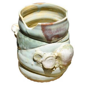 Puede incluir: Un jarrón de cerámica hecho a mano con un diseño texturizado único. El jarrón presenta una combinación de colores verde claro y marrón con una forma rugosa y orgánica. El borde superior tiene un borde roto, lo que aumenta su atractivo artístico.