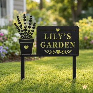 Könnte beinhalten: Ein schwarzes Metall-Gartenschild mit der Silhouette einer Lavendelpflanze im Topf auf der linken Seite und einer rechteckigen Tafel rechts. Die Tafel zeigt den Text LILY'S GARDEN in goldener Schrift, verziert mit einem Herz und Blattmotiven.