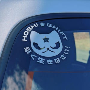 May include: A blue car window with a white sticker featuring a cartoon cat with a star on its head. The sticker says "HOSHI SHIFT" and "生きたない" in Japanese.