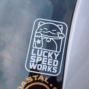 May include: White decal sticker with a cartoon cat holding a coin with a star on it. The text "Lucky Speed Works" is below the cat.