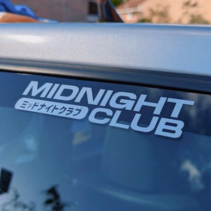May include: A silver car with a white sticker on the back window that says "MIDNIGHT CLUB" in bold white letters with the Japanese text "ミッドナイトクラブ" underneath.