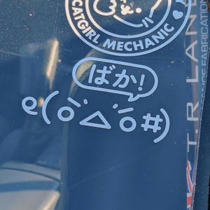 May include: A blue car window with a white sticker that says "e(o△o#)" and a white speech bubble sticker that says "ばか!" in Japanese. The car window also has a sticker that says "Catgirl Mechanic" and another sticker that says "T.R.L.A.N.E.  ICE FABRICATION"