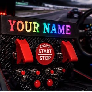 Op de afbeelding: Een zwarte button box voor racesimulatoren met een koolstofvezel textuur, voorzien van een centrale rode ronde engine start stop knop, twee grote rode tuimelschakelaars, vier kleinere metalen tuimelschakelaars en twee rode drukknoppen met regenboogverlichting.