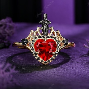 May include: A gold-toned ring featuring a heart-shaped red gemstone surrounded by black stones. A small sword design sits atop the heart. The ring has a crown-like design and the words "Dark Crown" are visible.
