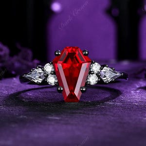 May include: A black ring with a coffin-shaped, deep red gemstone as the centerpiece. The ring is accented with white and clear stones on each side of the central stone. The background is a dark purple.