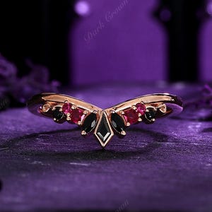 May include: A rose gold ring with a curved design, featuring a central black diamond-shaped stone, flanked by smaller red and black gemstones. The ring has a dark, elegant aesthetic.